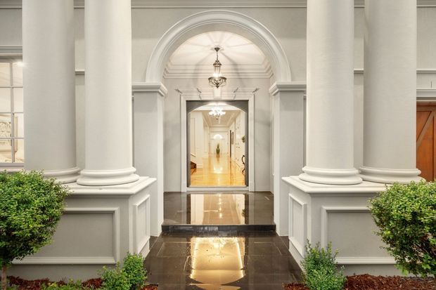 Formal classical-style entry facade with large white fluted columns, paneled bases, an arched portal, ornate pendant lights, and glossy dark stone paving leading into a bright interior corridor. Symmetrical clipped shrubs frame the entrance.