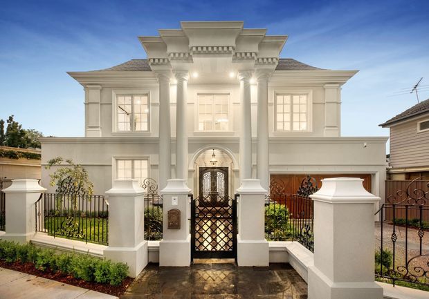 Front exterior view of a large, symmetrical two-storey residence with a pale rendered facade, elaborate classical columns and pilasters, ornate cornice detailing, hipped tiled roof, multi-pane windows, and an arched entry with decorative wrought-iron gates. A formal front garden, masonry fence posts, and paved path frame the entry.