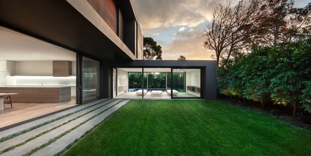 Contemporary two-storey residence viewed from a landscaped lawn and paved stepping-stone path. The dark, rectilinear facade features extensive floor-to-ceiling glazing, projecting upper-level volume and a sheltered courtyard-like living area. A warm, minimalist kitchen and interior spaces are visible through the open glazing, with mature planting surrounding the lawn.
