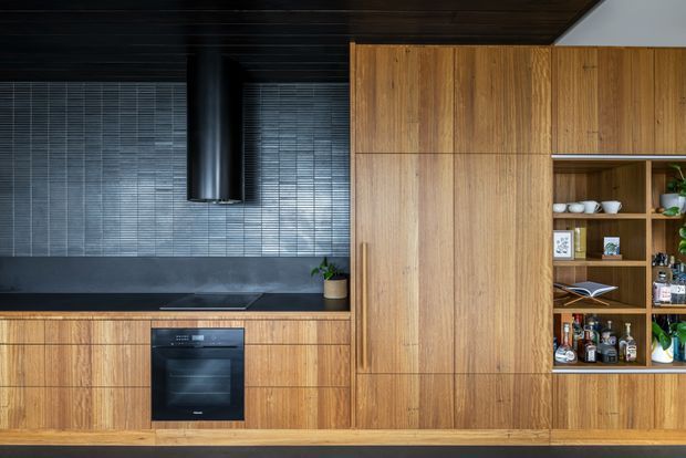 A contemporary galley-style kitchen featuring extensive warm timber cabinetry, a dark tiled splashback, integrated black appliances, open display shelving, and a dark timber ceiling. The composition emphasizes the built-in joinery and concealed storage, with a compact cooking zone and a bar-style shelving section containing glassware and bottles.