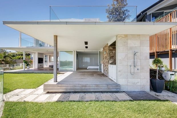 Bright exterior view of a contemporary multi-level residence with a broad cantilevered upper terrace, clear glass balustrades, timber decking, pale stone-clad walls, and an open ground-level outdoor living area. The covered space includes an outdoor shower and direct glazing into the interior, with landscaped lawn and stepping-stone paving in the foreground.