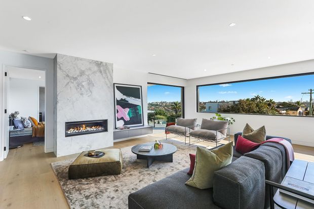 Bright contemporary living room with a large grey sectional sofa, sculptural coffee table, textured area rug, marble-look fireplace feature wall, built-in linear fire, and expansive horizontal windows overlooking a suburban neighbourhood. An adjacent room is visible through an open doorway.