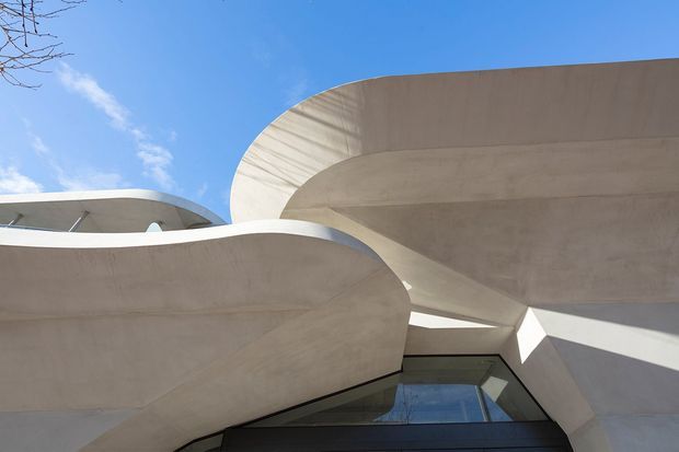 Upward-looking view of a sculptural contemporary building facade composed of sweeping curved concrete forms, deep overhangs, angular recesses, and a dark glazed opening. The pale concrete surfaces are strongly illuminated against a clear blue sky, creating a bold, abstract architectural composition.