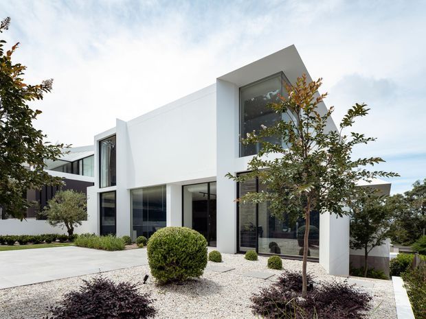 A contemporary multi-level white residential facade with strong geometric volumes, expansive dark-framed floor-to-ceiling glazing, and a projecting upper-storey form. The landscaped frontage features pale gravel, clipped rounded shrubs, small ornamental trees, and a broad light-paved driveway or entry court.