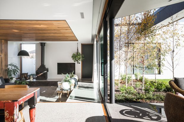Bright contemporary open-plan living space viewed from a glazed indoor-outdoor connection. The room features a warm timber-lined ceiling, dark-framed full-height glazing, a black freestanding wood-burning stove, television wall, light furnishings, and a rustic timber billiards table. A planted courtyard and adjacent building are visible beyond the glazing.