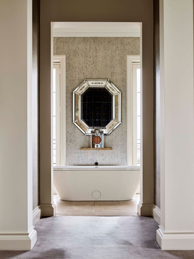 A symmetrical view through a carpeted hallway into an elegant bathroom. A freestanding white bathtub sits against a textured grey feature wall, beneath an ornate octagonal mirrored wall feature and a wall-mounted metallic tap. Tall narrow windows flank the bath, with substantial white moulded architraves, skirting boards and cornices. The room has a refined, traditional character with warm neutral tones and polished details.