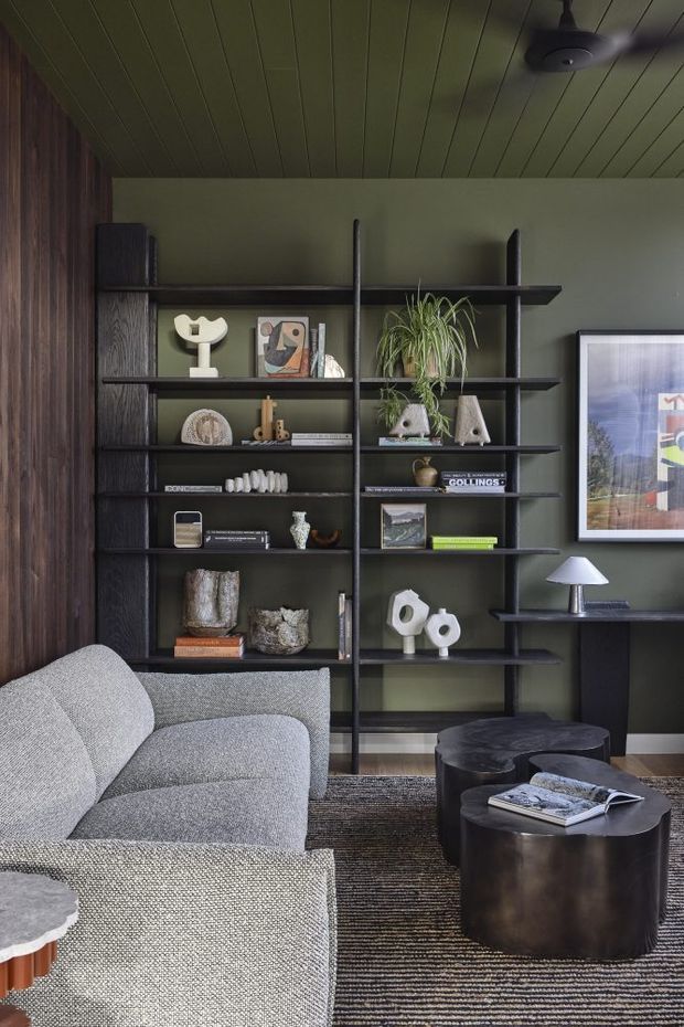 A moody, refined living room with a large built-in open shelving unit spanning the back wall. The shelves are dark-stained timber or dark-finished joinery with black vertical supports, styled with books, ceramics, framed artwork, and plants. A pale textured sectional sofa occupies the foreground, paired with dark rounded coffee tables and a striped area rug. The room has deep olive-green painted walls, a matching painted timber-plank ceiling, dark wood wall panelling at left, and a framed artwork above a slim dark console. A ceiling fan and table lamp complete the space.