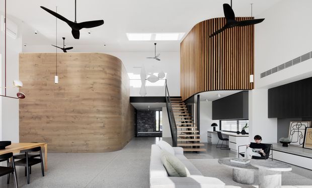 A bright, open-plan contemporary living space with a polished concrete floor, large curved timber-clad feature wall, sculptural timber mezzanine enclosure, and an open floating staircase. The room includes a white sofa, dining area, built-in media wall, and a compact workspace beneath the mezzanine. High ceilings, skylights, ceiling fans, and extensive open volume create an airy loft-like atmosphere.