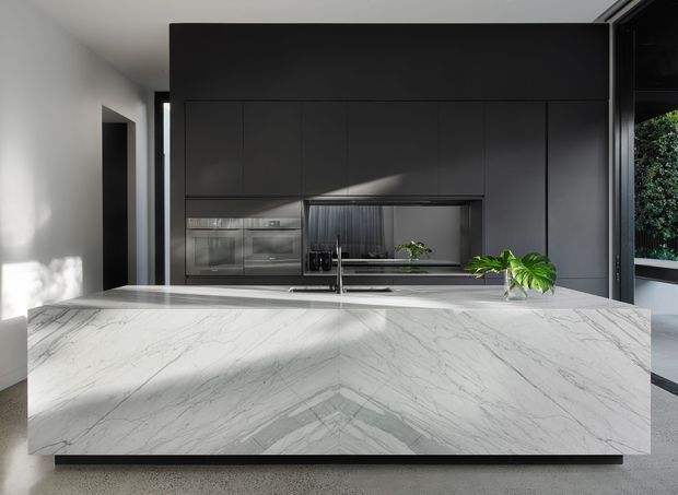 Contemporary kitchen featuring a large white veined stone island with a waterfall edge, dark full-height cabinetry, integrated stainless-steel appliances, a reflective splashback, and a broad window overlooking greenery. The space is clean-lined and strongly contrasted, with abundant natural light.