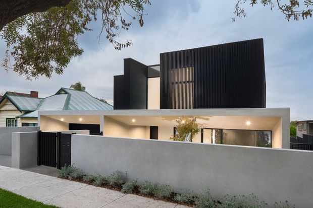 Street-facing view of a contemporary multi-level residence with a crisp rectilinear form. The home combines a pale rendered masonry boundary wall and covered entry/garage-like frontage with a dramatic upper volume clad in closely spaced dark vertical fins. Recessed warm lighting, slender dark window frames, and controlled landscaping create a refined modern facade. A neighbouring traditional house is visible behind the property.
