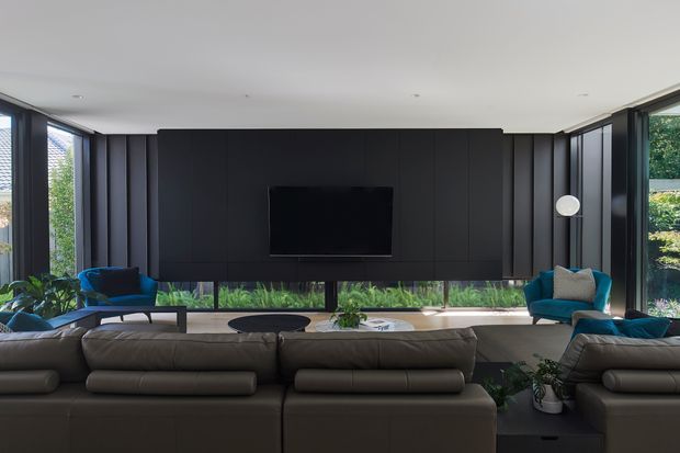 Contemporary living room with a large dark built-in media wall and wall-mounted television, flanked by vertical timber or dark-panel detailing and extensive floor-to-ceiling glazing. A large sectional sofa faces the television, with teal accent chairs, a low coffee table, indoor plants, and a bright landscaped outlook visible through the windows.