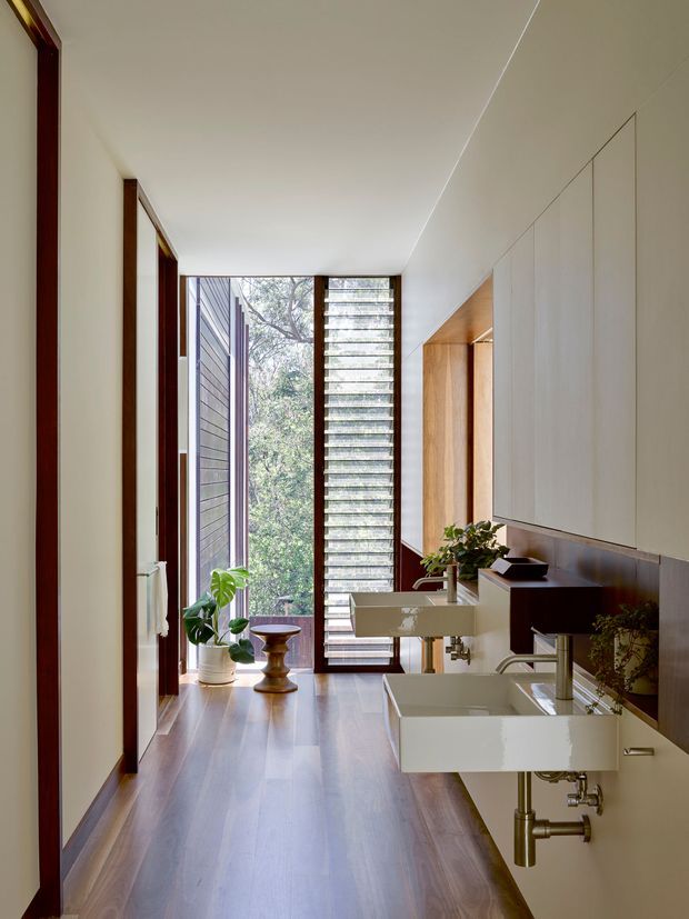 A narrow, bright contemporary bathroom with a long timber floor, double wall-mounted white ceramic basins, chrome wall-mounted taps, and extensive white built-in cabinetry. A tall louvred window and glazed opening at the far end bring in natural light and views of greenery, while dark timber door and window frames add contrast. Plants and minimal accessories soften the clean-lined space.