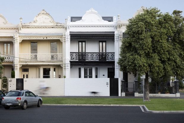 Street-facing view of a row of attached, ornate two-storey terrace houses. The central white facade has a decorative parapet, classical mouldings, black wrought-iron balcony railings, tall glazed doors and louvred shutters. Neighbouring cream and white heritage facades are visible, with a small front setback, lawn, mature tree and paved street.