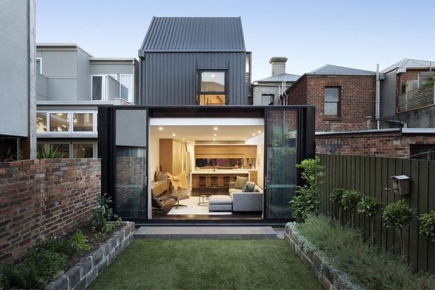 Rear exterior view of a compact contemporary urban residence, showing a dark corrugated-metal upper-level extension above a glazed ground-floor living and kitchen pavilion. The building opens onto a small landscaped courtyard with brick boundary walls, timber fencing, raised garden beds, and lawn. Neighbouring dense urban housing is visible around the property.