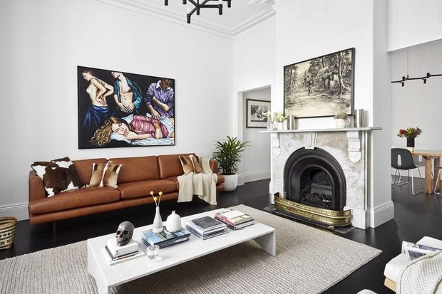 A refined living room in a period-style home, featuring a large cognac leather sofa, white walls with ornate ceiling cornices, dark timber flooring, a marble fireplace with a black cast-iron insert, and a contemporary white coffee table layered with books and decorative objects. Artwork and a glimpse of a dining area are visible beyond the room.