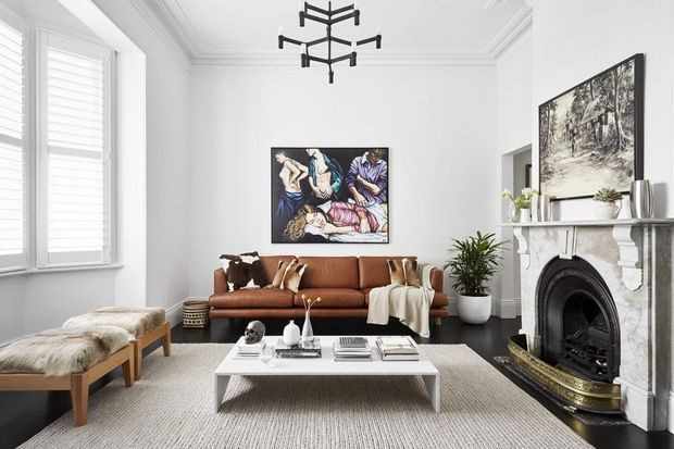 Bright, refined living room with white walls, decorative ceiling cornices, plantation shutters, a brown leather sofa, light upholstered stools, a low white rectangular coffee table, patterned rug, indoor plants, and an ornate marble fireplace with a black surround. A sculptural black pendant hangs centrally, while artwork and a large monochrome framed image add visual interest.