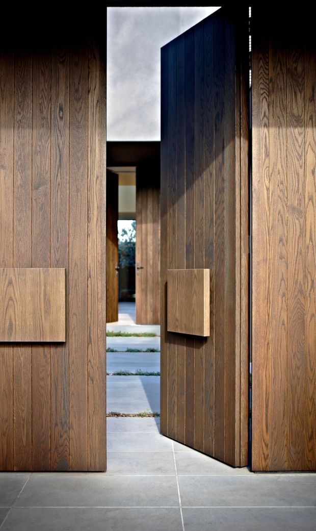 A dramatic close-up of a contemporary home entry featuring tall pivot doors and surrounding wall panels clad in vertically grained dark timber. The partially open door reveals a sheltered passage and stepped concrete paving beyond. Large-format grey concrete or stone tiles form the foreground floor, while strong sunlight creates deep shadows across the richly textured timber surfaces.