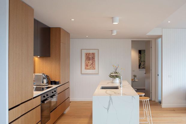 Bright contemporary kitchen with a long central island, integrated timber cabinetry, white marble-look benchtop, timber flooring, and vertically panelled white walls. The kitchen includes a built-in oven and cooktop, concealed rangehood, small appliances, pendant-free recessed lighting, and stools at the island. A doorway leads to an adjacent space in the background.