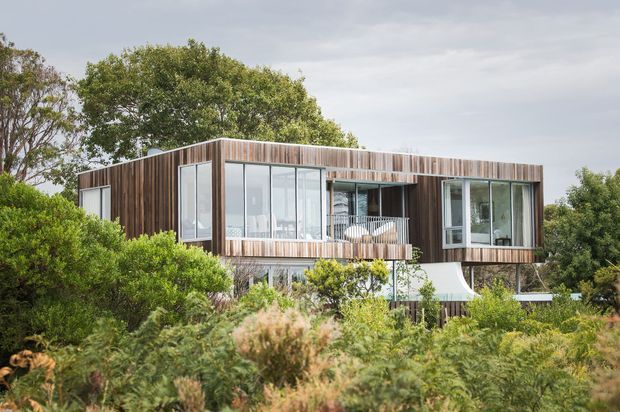 Exterior view of a contemporary elevated house set within dense native landscaping. The building has a simple box-like form with extensive glazing, vertical timber cladding, a projecting balcony, and a partially visible curved white structural element beneath the upper level.