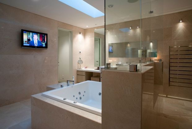 Luxurious contemporary bathroom with a large built-in spa bath, glass-enclosed shower, wall-mounted television, double vanity, extensive mirrors, recessed lighting, and warm beige stone finishes. A skylight brings natural light into the otherwise refined, hotel-like space.