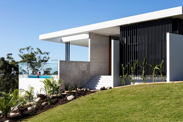 A contemporary flat-roofed house on a gently sloping landscaped site, featuring crisp white roof planes, exposed concrete walls, broad outdoor steps, slim dark vertical screening, extensive glazing, and a partially visible swimming pool. Tropical planting and a bright blue sky surround the minimalist exterior.