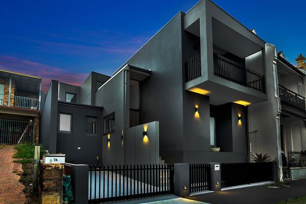 Twilight exterior view of a narrow, multi-level contemporary townhouse or residential extension. The dark rendered facade features strong geometric volumes, recessed balconies, black metal balustrades and fencing, compact windows, and warm integrated wall lighting. Adjacent traditional terrace-style buildings are visible on either side, with a paved driveway and front gate in the foreground.