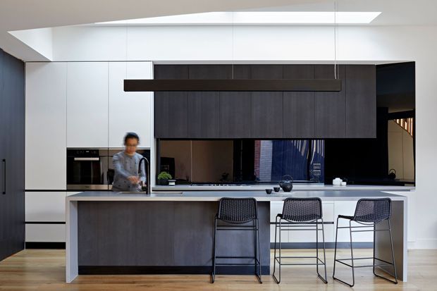 A contemporary open-plan kitchen dominated by a long dark timber-toned island with three bar stools. Full-height white cabinetry and integrated appliances line the rear wall, while dark overhead cabinetry frames a black reflective splashback. A linear pendant light hangs above the island, with a skylight bringing in natural light. Timber flooring and a glimpse of a stairway are visible.