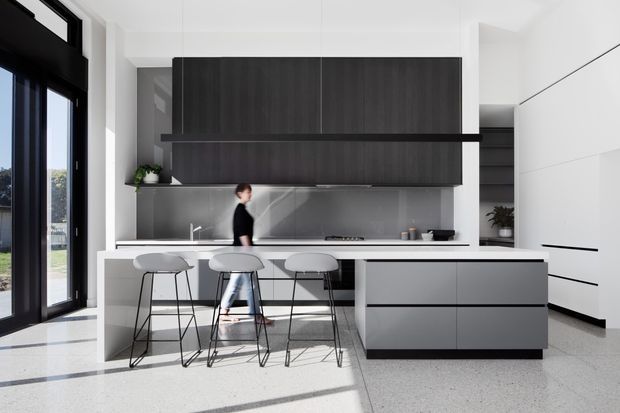 A bright, contemporary kitchen with a long island and three slim bar stools. The space features dark timber-look overhead cabinetry, a pale stone or solid-surface benchtop, a reflective grey splashback, white walls and full-height black-framed glazing. Integrated white cabinetry and strong natural light create a clean, minimalist composition.
