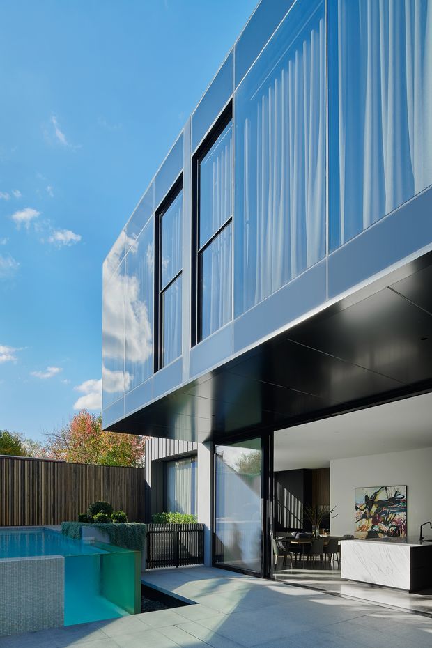 Contemporary multi-storey residential exterior with a projecting upper level clad in reflective silver panels and tall vertical glazing screened by sheer curtains. The ground floor opens onto a paved pool terrace, revealing a bright kitchen and dining area with dark-framed glazing, dark timber wall panelling, and a white stone island. A dark privacy fence and landscaped pool edge frame the outdoor space.