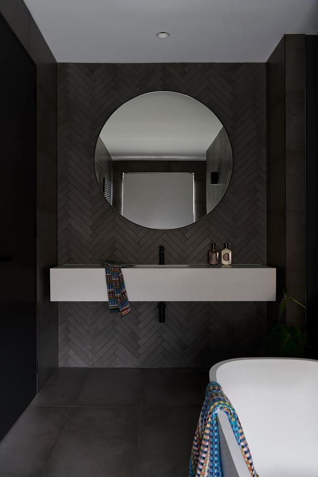 A dark, refined bathroom featuring a wall-mounted white vanity, large round mirror, black wall-mounted tapware, and a freestanding white bathtub. The feature wall is clad in dark herringbone-patterned tiles, complemented by large dark floor tiles and dark cabinetry. A colourful patterned textile adds a small accent of colour.