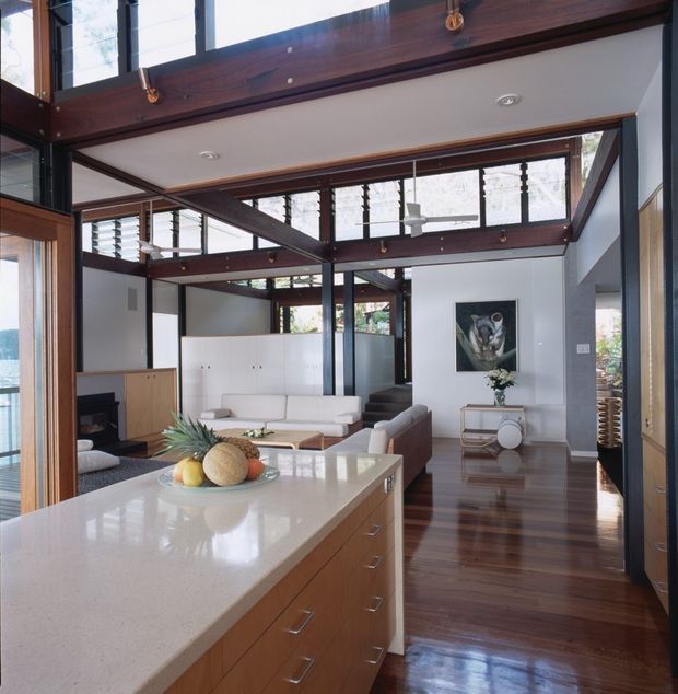 A bright, open-plan living and kitchen interior with a large island bench in the foreground, white integrated cabinetry, timber drawer fronts, polished light benchtop, and warm dark polished timber flooring. The living area includes white sofas, a fireplace, a glass coffee table, and a small console. Extensive clerestory glazing and dark timber structural beams create a lofty, light-filled space with strong indoor-outdoor connection and exposed post-and-beam detailing.