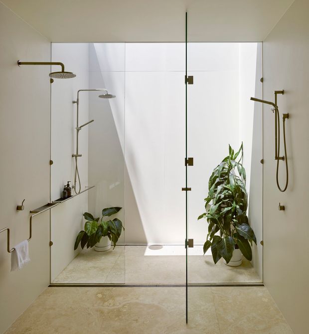 Bright contemporary bathroom with a large frameless glass shower enclosure, multiple wall-mounted brass shower fixtures, pale stone or porcelain wall and floor finishes, and integrated niches and ledges. Several potted tropical plants soften the minimalist space, while a concealed skylight or high-level opening creates strong natural light and shadows.