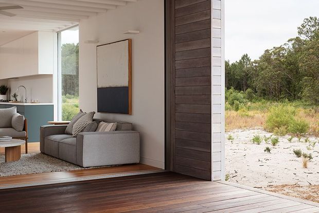 A contemporary indoor-outdoor living space with wide-open glazed or folding access to a timber deck. The interior contains a grey sectional sofa, armchair, artwork, and a compact kitchen with muted blue-green cabinetry. White walls and exposed painted ceiling beams contrast with dark timber wall cladding and decking, while a natural bush landscape is visible outside.