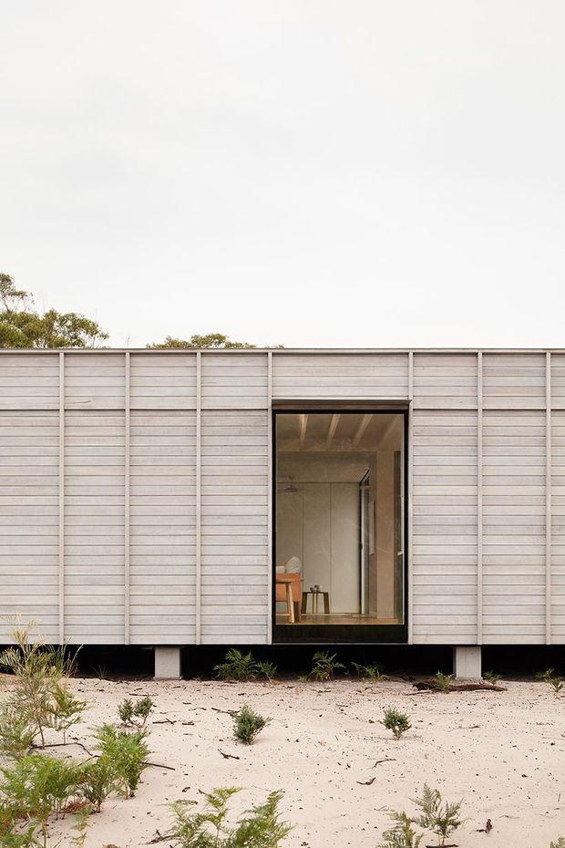 Exterior view of a compact elevated building with a restrained modern facade clad in pale horizontal boards. A large central glazed opening reveals a simple interior, while the structure is supported on short piers above sandy, lightly vegetated ground. The composition emphasizes the horizontal cladding, dark recessed opening, and minimal low-impact setting.