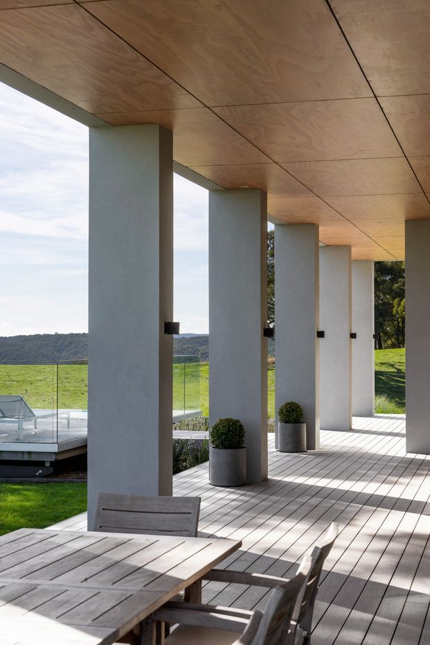A covered outdoor terrace with a long timber-lined ceiling, pale rendered structural columns, and timber decking. A timber dining table and chairs occupy the foreground, while glass balustrades, potted topiary, a lawn, and a distant wooded hillside create an open landscaped setting. The design is clean and contemporary, with strong linear perspective and abundant daylight.