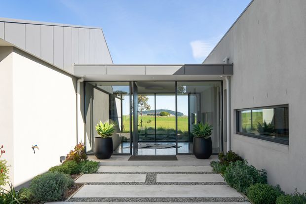 A modern residential entrance courtyard framed by clean, angular volumes and extensive full-height glazing. A covered glass entry links the two pale rendered wings, with dark metal window and door frames, large black planters, and a structured pathway of pale concrete slabs set in fine gravel. Rolling green rural landscape is visible beyond the glazed entry.