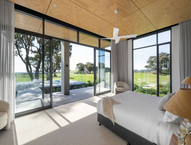 A bright contemporary bedroom with a large bed positioned beside expansive black-framed floor-to-ceiling glazing. The room features a warm plywood-lined ceiling, pale carpet, full-height grey curtains, a white sculptural ceiling fan, and a small upholstered lounge chair. Glazing opens toward a covered outdoor deck and expansive green rural landscape, with strong sunlight casting geometric shadows across the floor.
