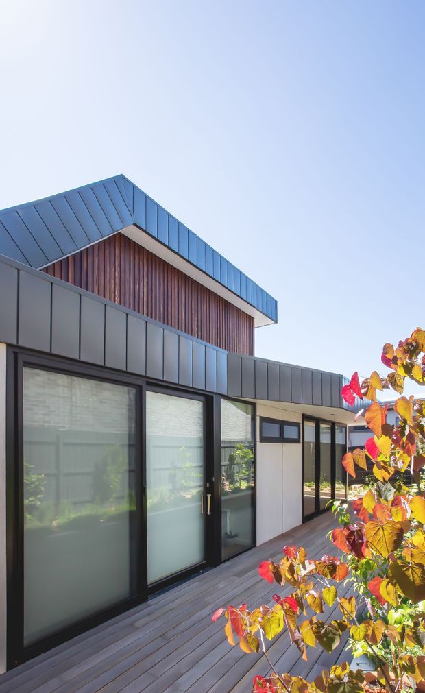 Exterior view of a contemporary single-storey dwelling with a raised gabled upper volume. The facade combines dark metal cladding, extensive black-framed glazing, and vertical timber wall cladding, opening onto a timber deck. Autumn foliage occupies the foreground under a clear sky.