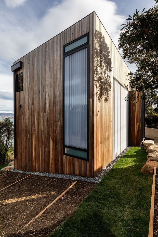 Exterior view of a compact modern dwelling or extension with a sharply pitched asymmetrical roofline. The facade is clad in vertical natural timber boards, with contrasting dark-framed tall translucent glazing and a secondary light-coloured vertical cladding section. The building sits on a landscaped sloping site with gravel edging, lawn, rocks, and mature vegetation; a distant coastal or water view is visible at the left.