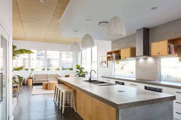 Bright open-plan kitchen with a large timber island, grey stone benchtop, integrated sink and seating, opening into a small living area. The kitchen features light timber cabinetry, white lower cabinets, a pale textured tile splashback, open shelving, and woven pendant lights. Large windows provide abundant natural light, while a timber-lined ceiling panel adds warmth.