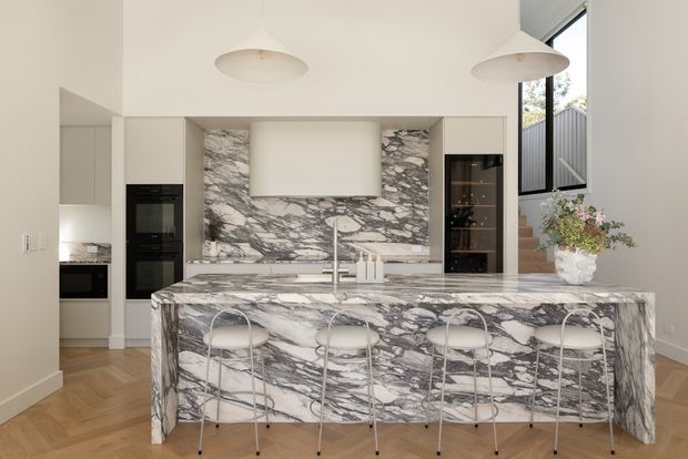 Bright, refined contemporary kitchen with a large waterfall island clad in dramatic grey-and-white veined stone. The island seats four on slim metal-framed stools and faces a matching stone splashback behind the cooktop and sink area. Light neutral cabinetry, integrated appliances, open shelving, glass-front display storage, pendant lights, timber parquet flooring, and a stairway with tall glazing are visible.