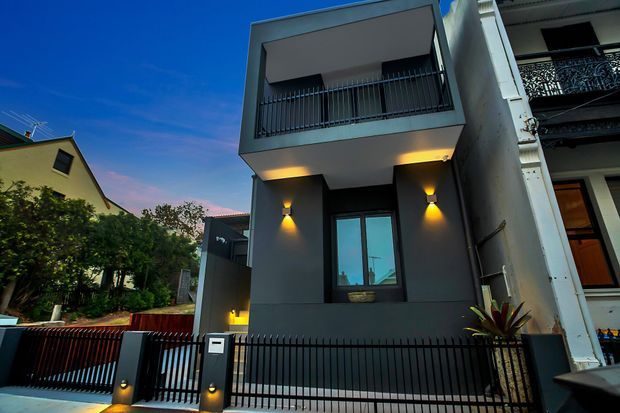 Street-facing view of a narrow, multi-storey contemporary townhouse with a dark grey rendered facade, flat roofline, projecting upper-level balcony, black metal balustrades and fencing, and warm wall-mounted exterior lighting. Adjacent residential buildings and landscaping are visible on either side.