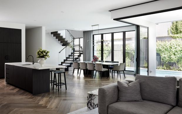 Bright open-plan contemporary home interior with a dark kitchen island in the foreground, dining area, floating staircase with black metal balustrade, and large floor-to-ceiling glazed doors opening toward a garden and pool. A grey sectional sofa occupies the living area, and herringbone timber flooring runs throughout the space.