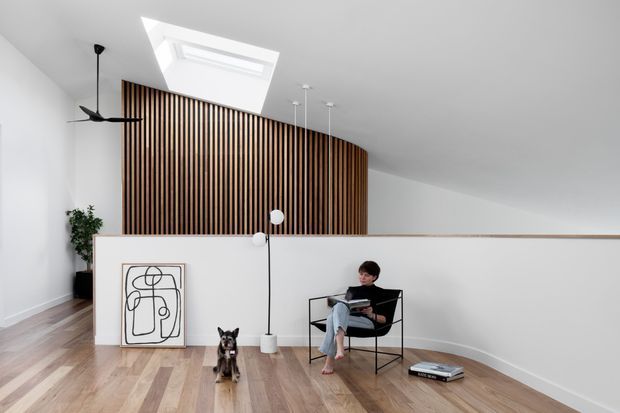 Bright contemporary interior with a curved vertical timber slat feature wall forming a partial divider, white walls and ceiling, a skylight, light timber flooring, and minimalist black furniture. A person is seated in a lounge chair beside a sculptural floor lamp, with artwork and a small dog in the foreground.