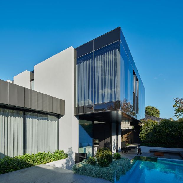 A contemporary multi-level residential facade with crisp white rendered walls, dark metal cladding, extensive floor-to-ceiling glazing, and projecting upper volumes. The building overlooks a landscaped courtyard and rectangular swimming pool, with sheer curtains visible behind the glazing and a sheltered outdoor seating area beneath the cantilevered upper storey.