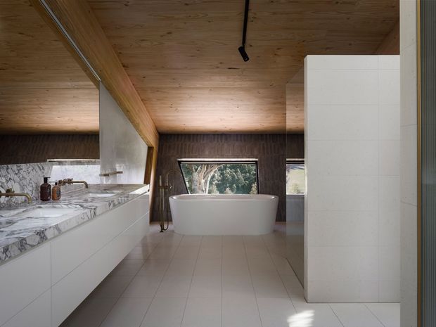 A spacious contemporary bathroom with a freestanding white bathtub positioned in front of a large window, a long white vanity with a dramatic grey-veined stone benchtop, and pale tiled flooring. The room features warm timber ceiling lining and exposed timber beams, dark textured wall finishes, minimalist fixtures, and views of trees through the glazing.