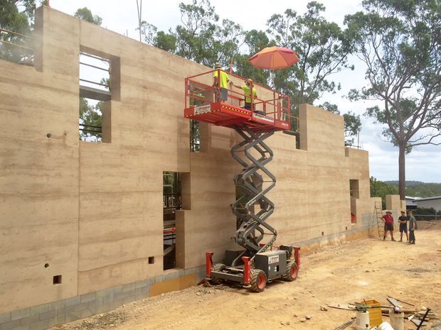 An exterior construction-site photograph showing a partially built modern concrete house with large rectangular openings and tall solid wall planes. Workers are using a red scissor lift against the facade, with construction materials and equipment in the foreground. Eucalyptus trees and a suburban landscape are visible behind the building.