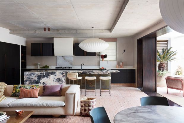 Open-plan contemporary kitchen and living area with a dramatic veined stone island and matching round dining table, dark timber cabinetry, pale stone or plaster walls, and an exposed concrete ceiling. A cream sofa with colourful cushions faces the kitchen, while sculptural pendant lights, bar stools, and an indoor potted palm add a refined, eclectic character. Large openings connect the interior to a bright outdoor area.
