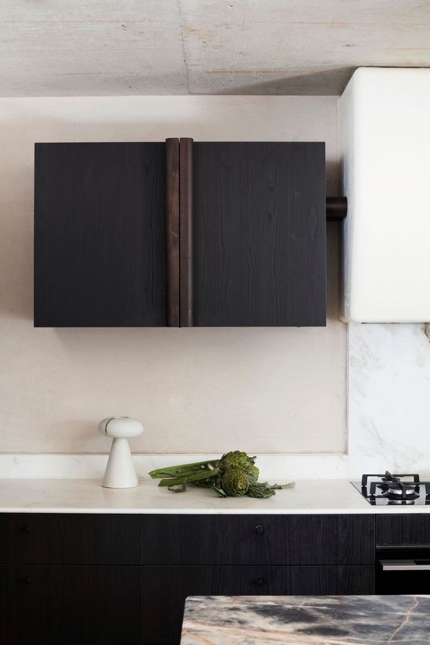 Close-up view of a refined contemporary kitchen, featuring dark vertically grained timber cabinetry, a pale stone benchtop and splashback, a compact gas cooktop, and a sculptural white countertop lamp. A dark wall-mounted cabinet and marble-look island benchtop add strong contrast, while the exposed pale concrete ceiling introduces an understated industrial character.