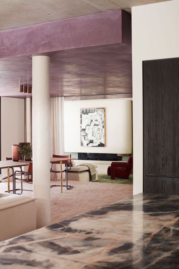 A refined open-plan residential interior viewed from behind a dark marble kitchen or bar benchtop. The space includes a dining area with a timber table and chairs, a living area with a sofa, red lounge chairs, television and artwork, pale curtains, a cylindrical column, and a dramatic mauve-toned ceiling and beam. The palette combines blush pink, cream, burgundy, dark timber and richly veined marble, creating a sophisticated, contemporary atmosphere.